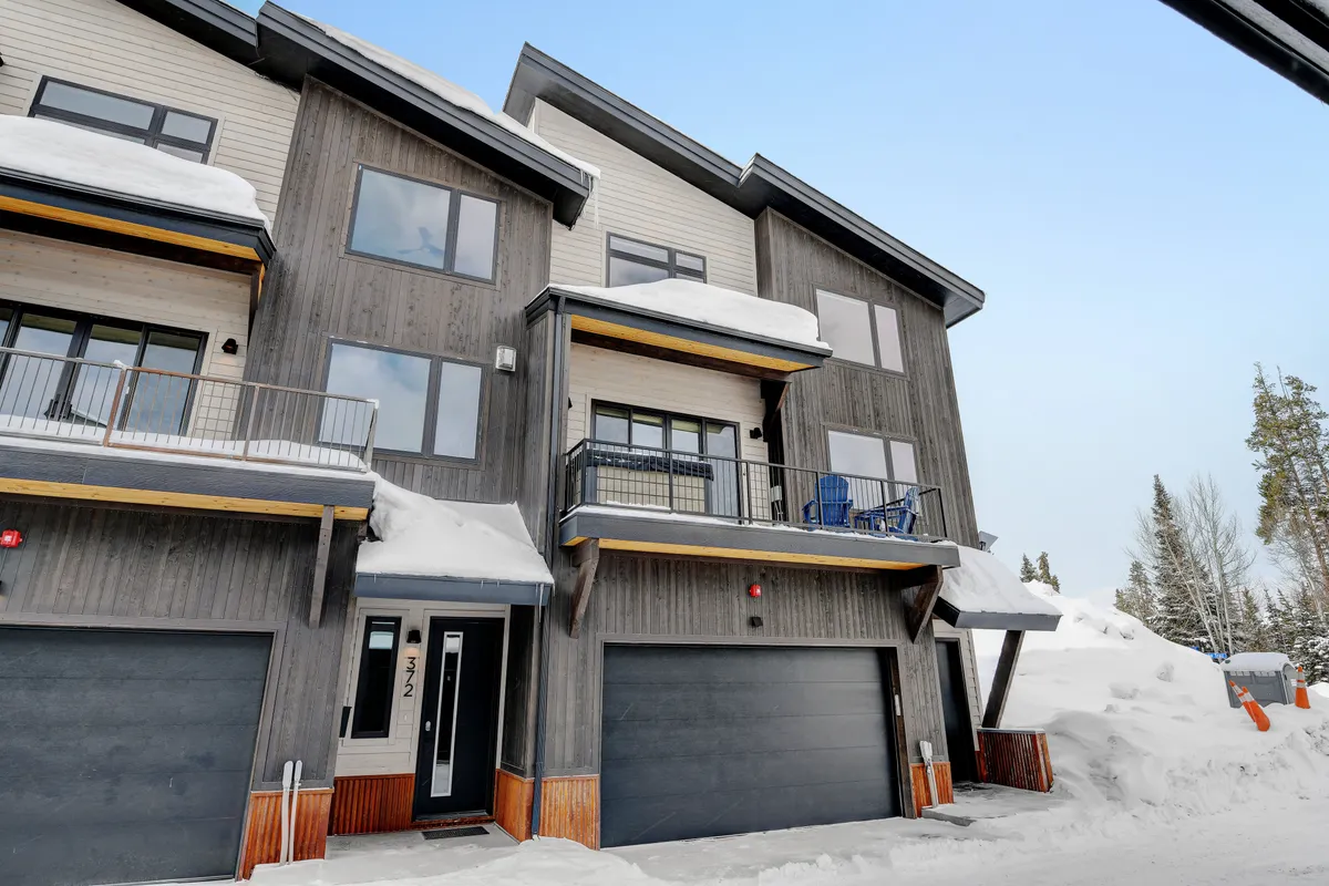 A welcoming exterior view of the mountain home featuring a wooden facade, spacious balconies, expansive windows, and garage doors. Snow lightly covers the scene, evoking a sense of winter retreat and group gatherings.