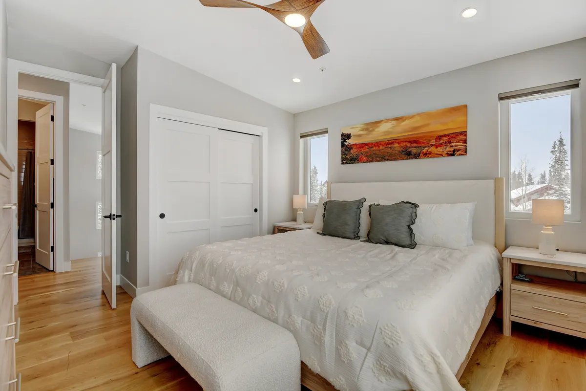A beautifully made bed adorned with decorative pillows sits between matching nightstands, each with its own lamp. Soft artwork hangs above the headboard, and natural light streams in through a nearby window, creating a serene and cozy bedroom atmosphere.