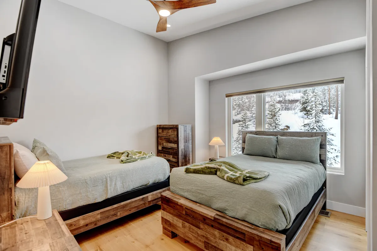 A spacious bunk room featuring two full beds with cozy linens, a large window allowing in serene mountain views, and a nightstand with a lamp nestled between the beds. The room exudes calmness, perfect for teens, friends, or children seeking their own space.
