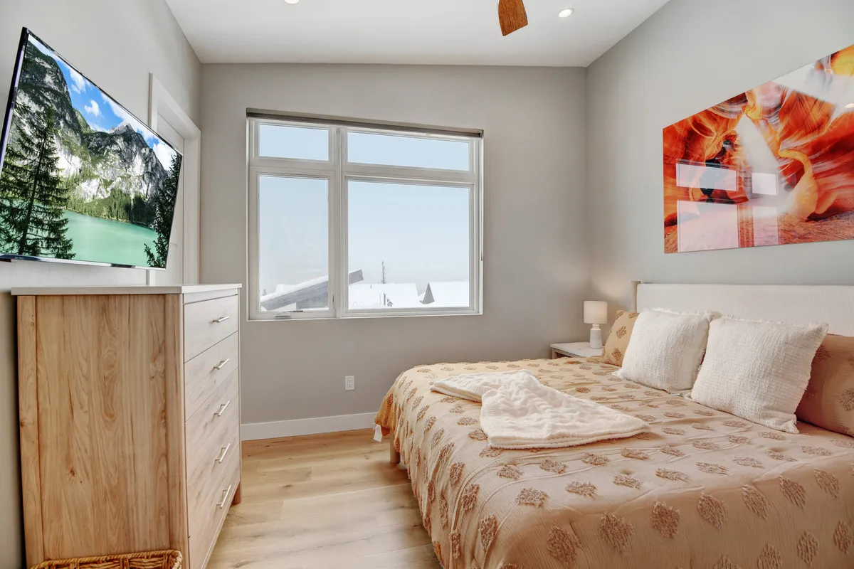 A cozy bedroom setup featuring a bed with a decorative blanket, a large TV placed on a dresser, artwork on the wall, and natural light streaming through a window. The room invites guests to enjoy both restful sleep and relaxing movie nights in privacy.