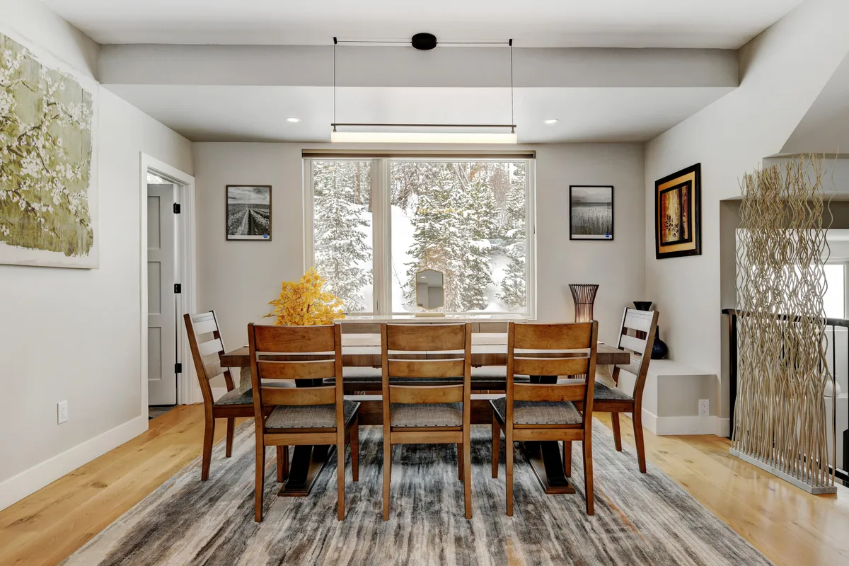 A dining area with a spacious table is set next to a large window overlooking a snowy mountain scene. Subtle décor, a decorative plant centerpiece, and tasteful wall art contribute to a warm and cozy ambiance ideal for family meals or game nights.