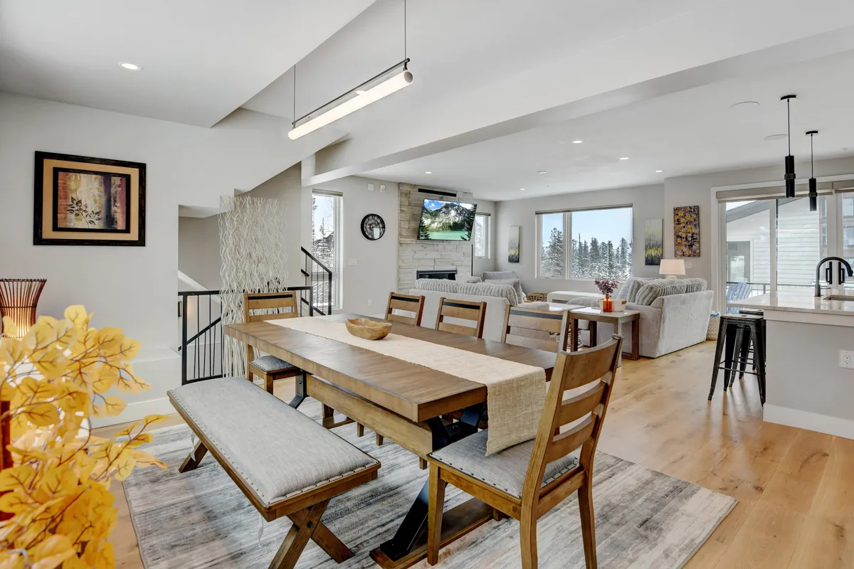 The image showcases a spacious, warmly lit living and dining area with a cozy fireplace, a comfortable sofa arrangement, and a large dining table perfect for group meals. Expansive windows reveal sweeping mountain views, filling the space with natural light and connecting the indoors to the scenic Colorado landscape.