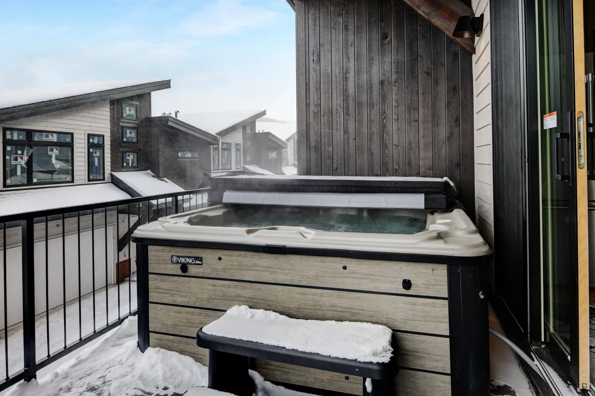 A serene outdoor hot tub set on a snow-covered balcony, surrounded by mountain architecture and neighboring buildings. The hot tub offers a luxurious place to unwind after a day of outdoor adventure, with tranquil winter views enhancing the ambiance.