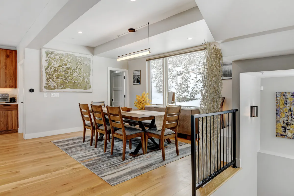 A large dining table sits in a warm, light-filled space, surrounded by modern decor and large windows. The table is set for a family or group, emphasizing the communal aspect of dining together.