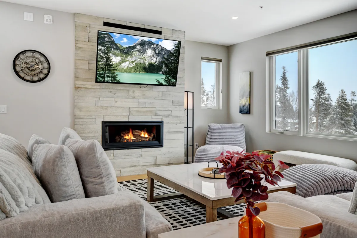 A warm, open living room features a cozy sofa arranged around a modern gas fireplace and a wall-mounted TV, with expansive windows offering breathtaking mountain views. The coffee table invites group activities, and the overall space is designed for both relaxation and social gatherings.