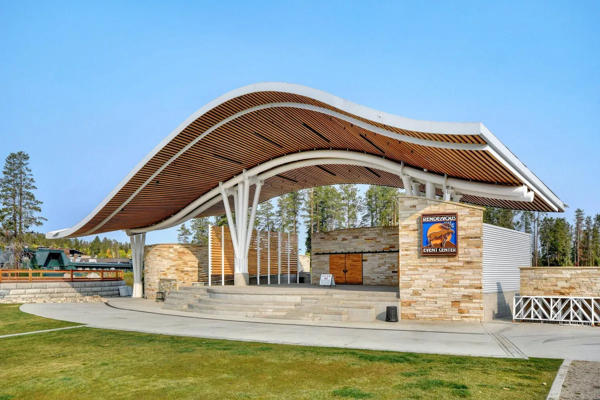 An outdoor, community gathering space with a dramatic curved roof structure, stone facade, and open stage area, set up for events and local celebrations. The festive atmosphere, with people gathered for performances, highlights communal and celebratory activities.