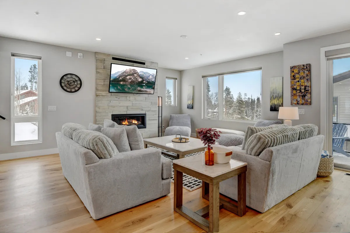 A spacious living room showcases a stylish seating arrangement beneath decorative wall art, with a fireplace and television set against a backdrop of floor-to-ceiling windows. Daylight fills the space, emphasizing a welcoming atmosphere suited for family gatherings or quiet moments.