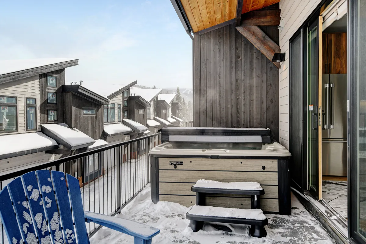 An exterior view of a hot tub positioned on a snow-covered balcony with a blue chair nearby, all set against a tranquil winter backdrop. The scene evokes relaxation, with the promise of soaking in the hot tub while enjoying serene mountain views.
