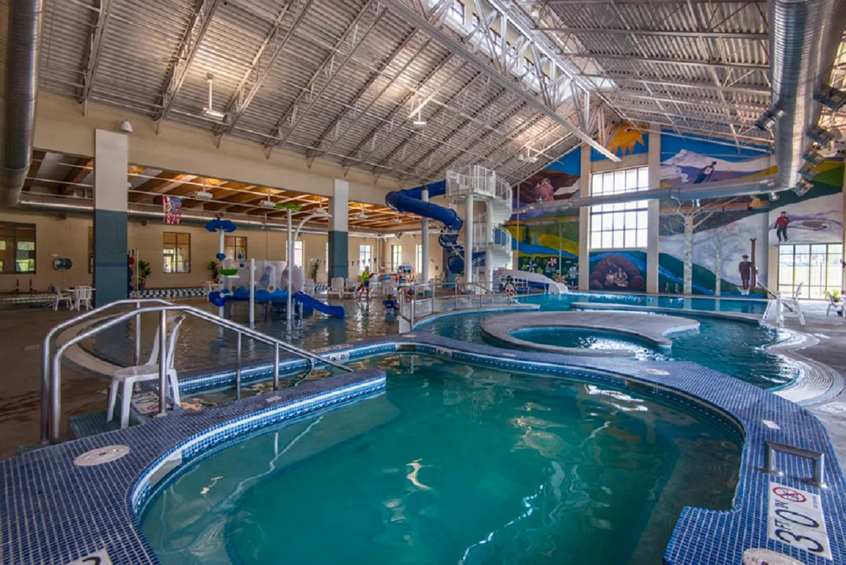 A lively indoor recreation center with a large pool, water slides, a mural, and children playing and socializing, with parents relaxing on lounge chairs nearby.