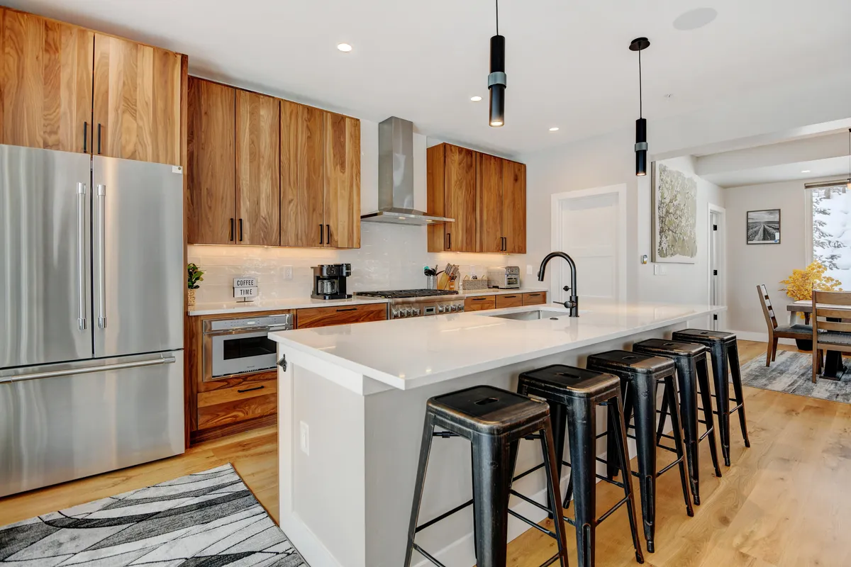 This image features a modern kitchen with a prominent island, sleek stainless steel appliances, wooden cabinetry, bar stools for casual seating, and a dining area in view. The setup highlights ample counter space and an inviting atmosphere for gatherings.