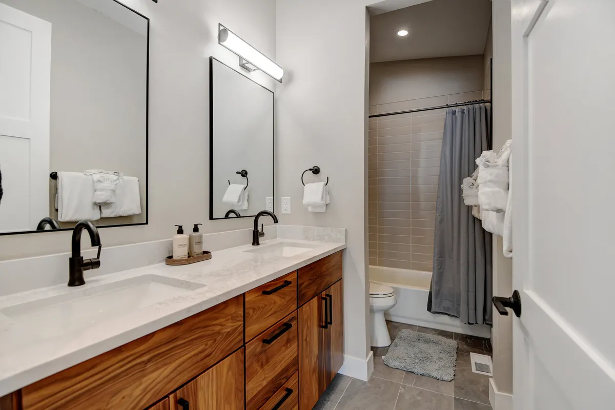An elegantly appointed bathroom featuring a double sink vanity, generously sized mirrors, wooden cabinetry, and a shower area. Morning light reflects off the surfaces, creating a tranquil and inviting space perfect for self-care and relaxation.