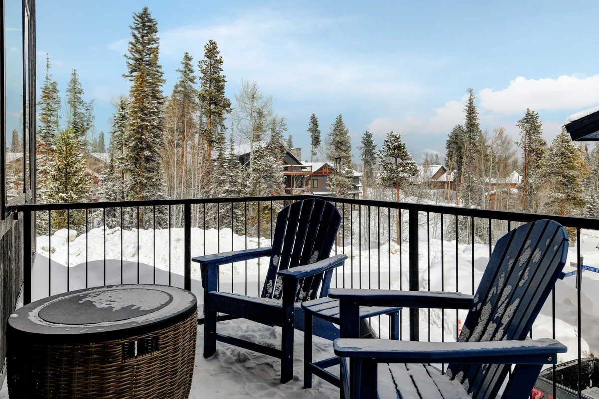 A patio scene with blue Adirondack chairs on a snow-covered ground, framed by distant pine trees and neighboring cabins. The setting highlights a peaceful spot for outdoor lounging, enjoying mountain views, and embracing seasonal changes.